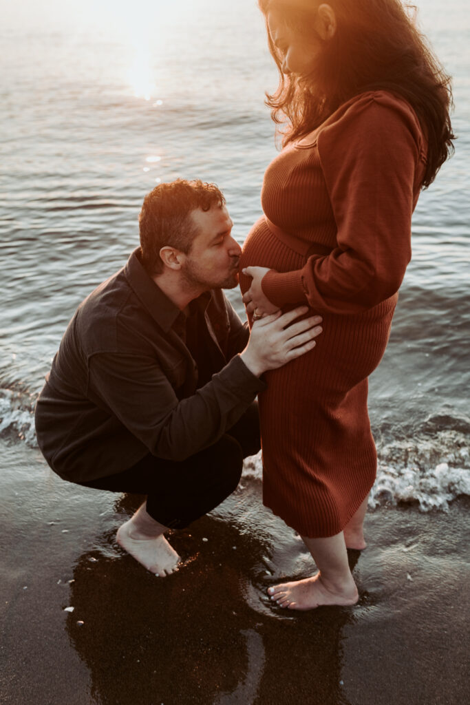 Expecting couple on a Vancouver beach, partner kneeling by the bump